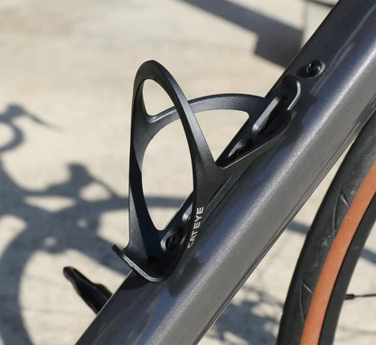 Close-up of a black CATEYE BC-400 Bottle Cage mounted on a bicycle frame, with a blurred background and the bike’s shadow visible on the ground.
