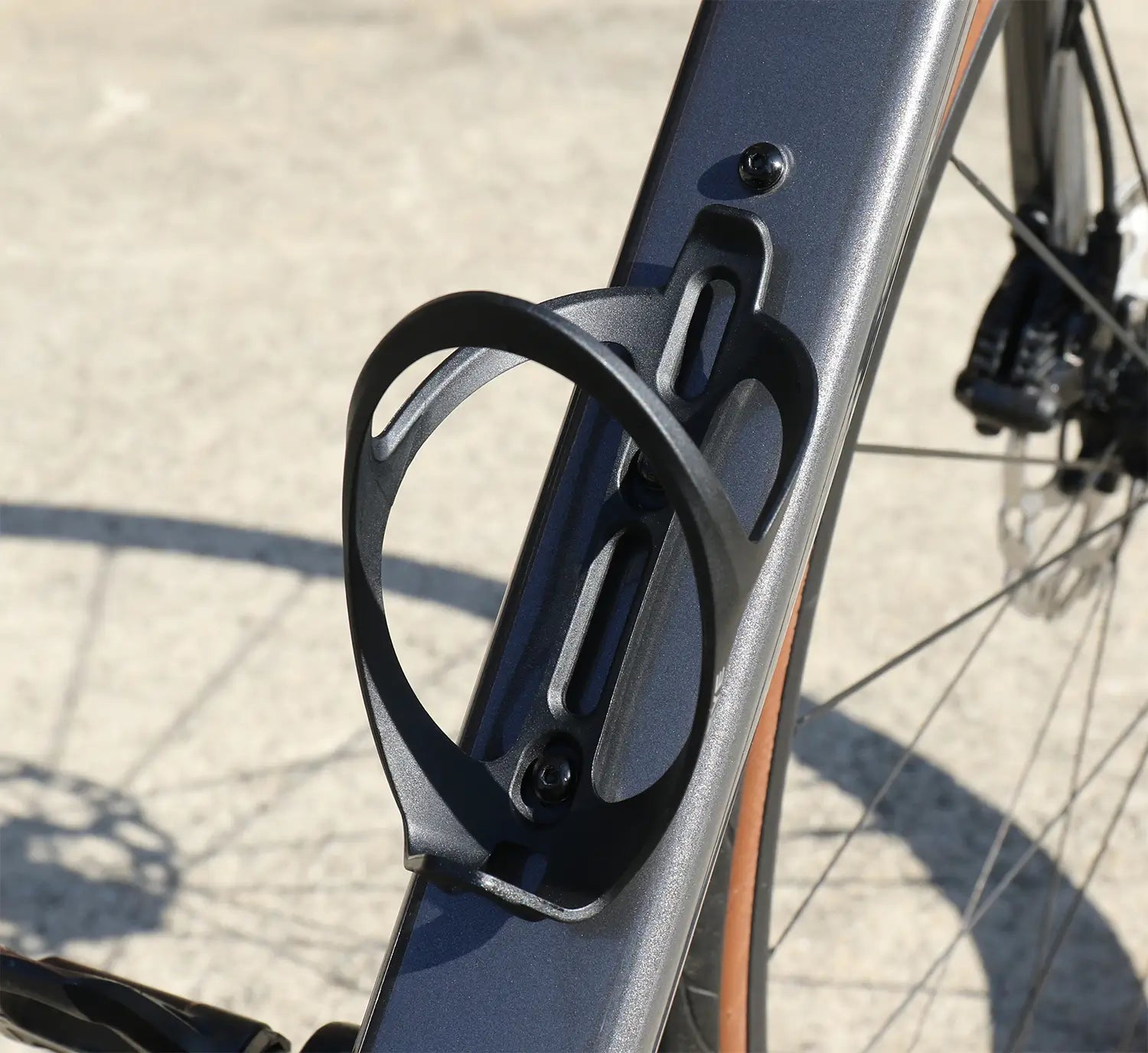Close-up of the CATEYE BC-400 Bottle Cage mounted on a metallic bike frame, with the rear wheel and disc brake in view. Shown outdoors on a light surface, it highlights the cage’s universal bike compatibility.