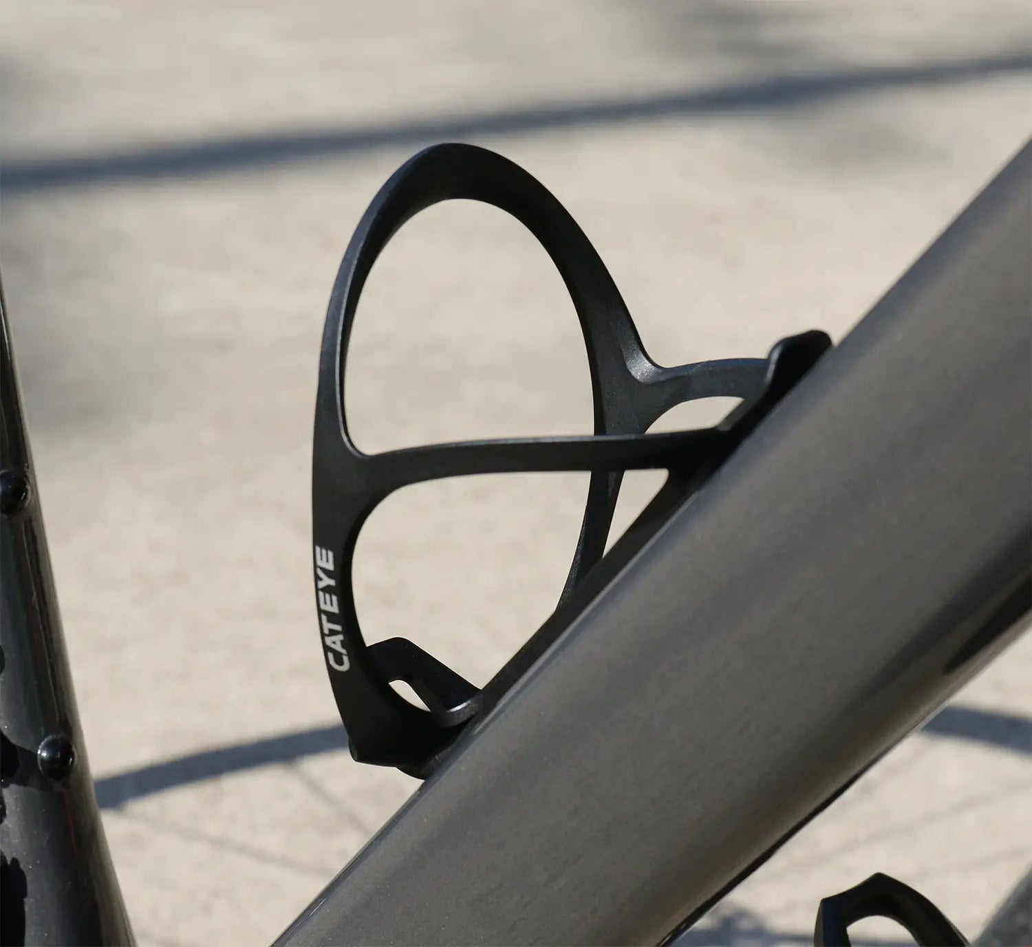 Close-up of a CATEYE BC-400 Bottle Cage in black, mounted on the frame of a gray bike and photographed outdoors in sunlight.