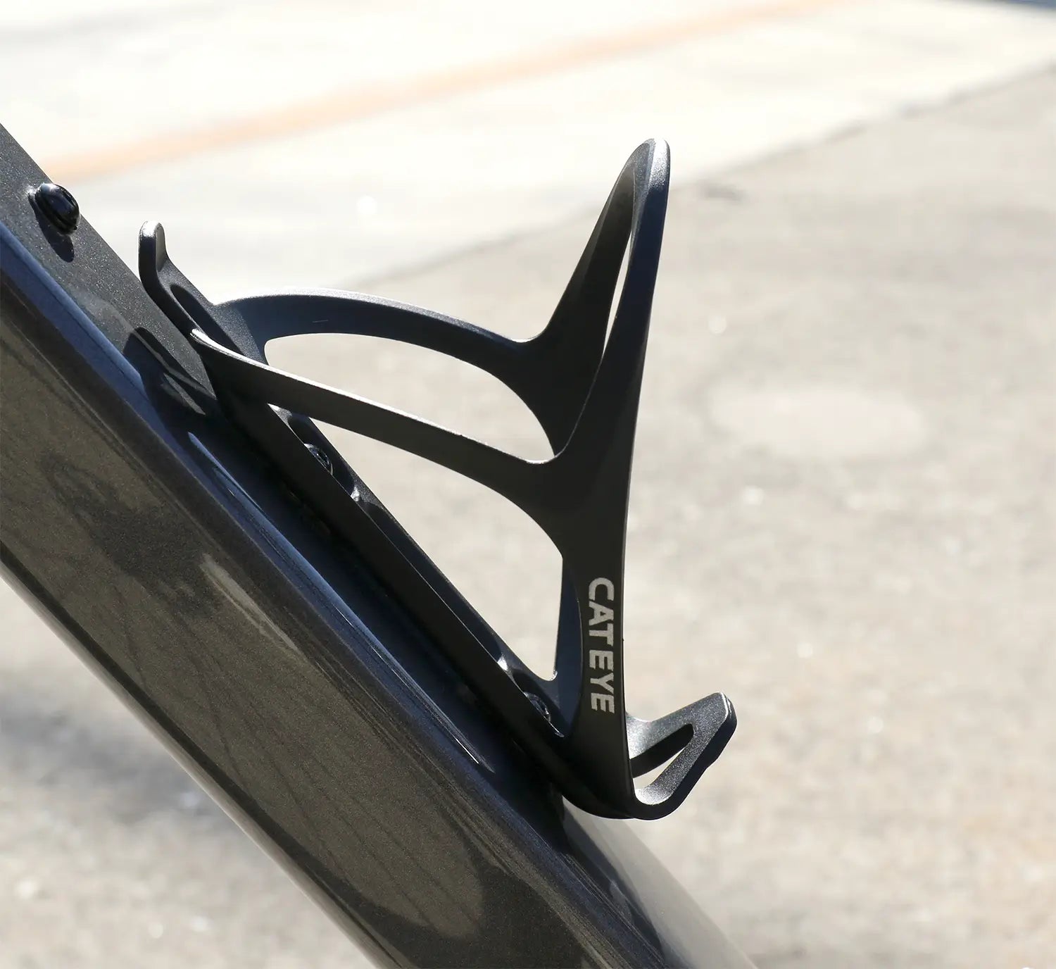 A CATEYE BC-400 Bottle Cage in black is attached to the down tube of a dark bike frame, outdoors on a sunny day.