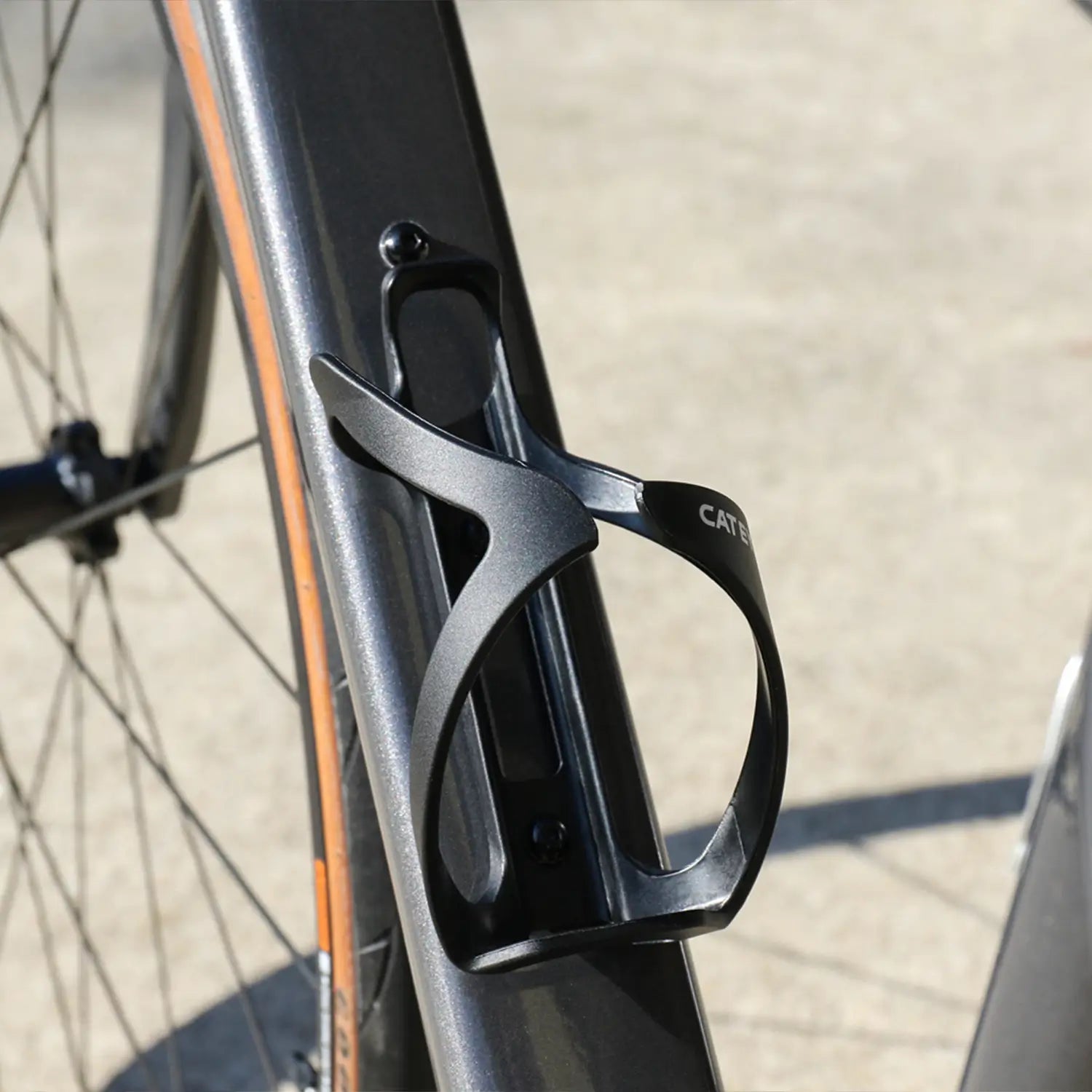 A close-up of the CATEYE BC-500 Bottle Cage, a black side-loading design, mounted on a bike frame with part of the rear wheel visible. The sunlit surface and blurred background suggest an adventure-ready ride.
