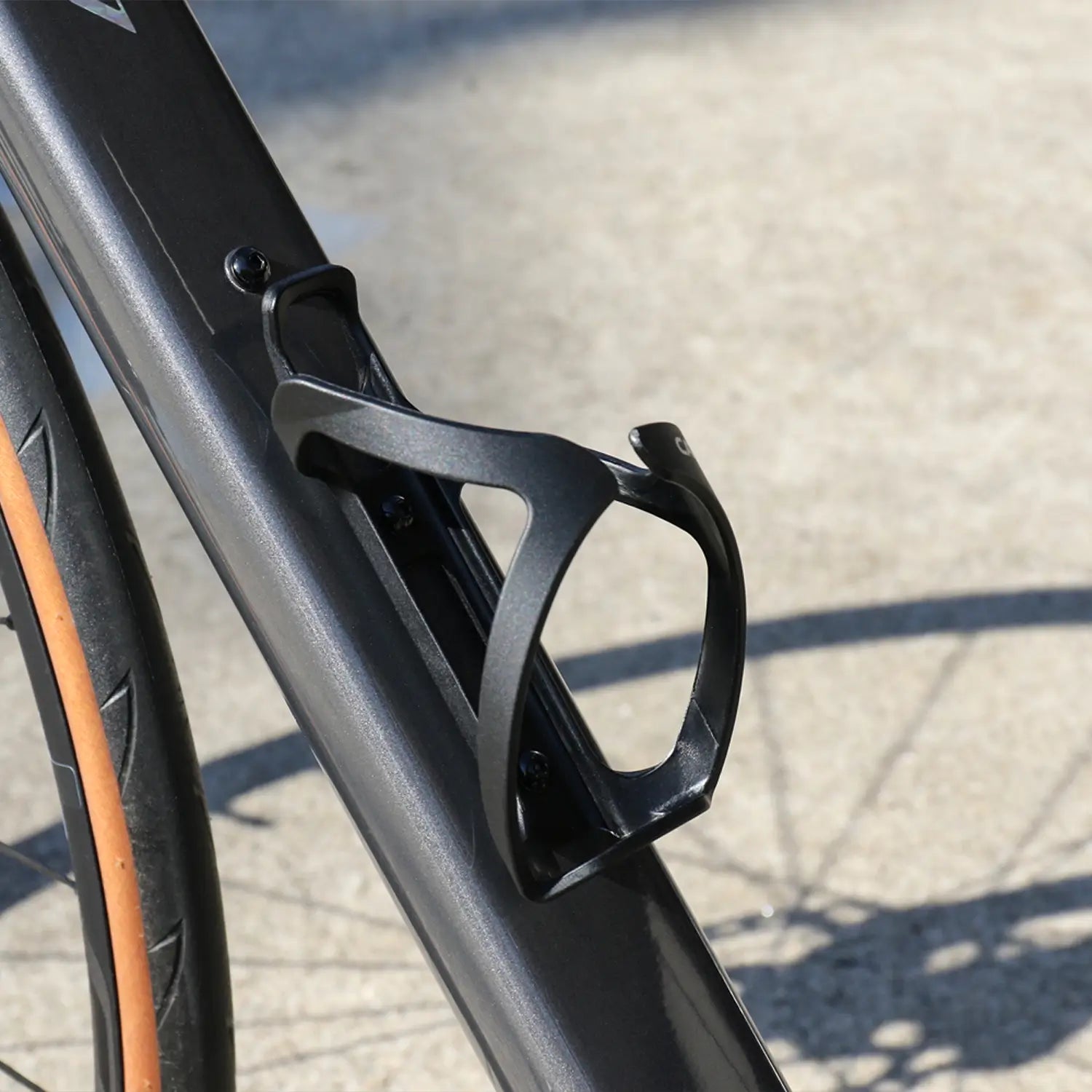 A close-up of the CATEYE BC-500 Bottle Cage in black, mounted on a bike frame, with sunlight casting shadows on the pavement below.