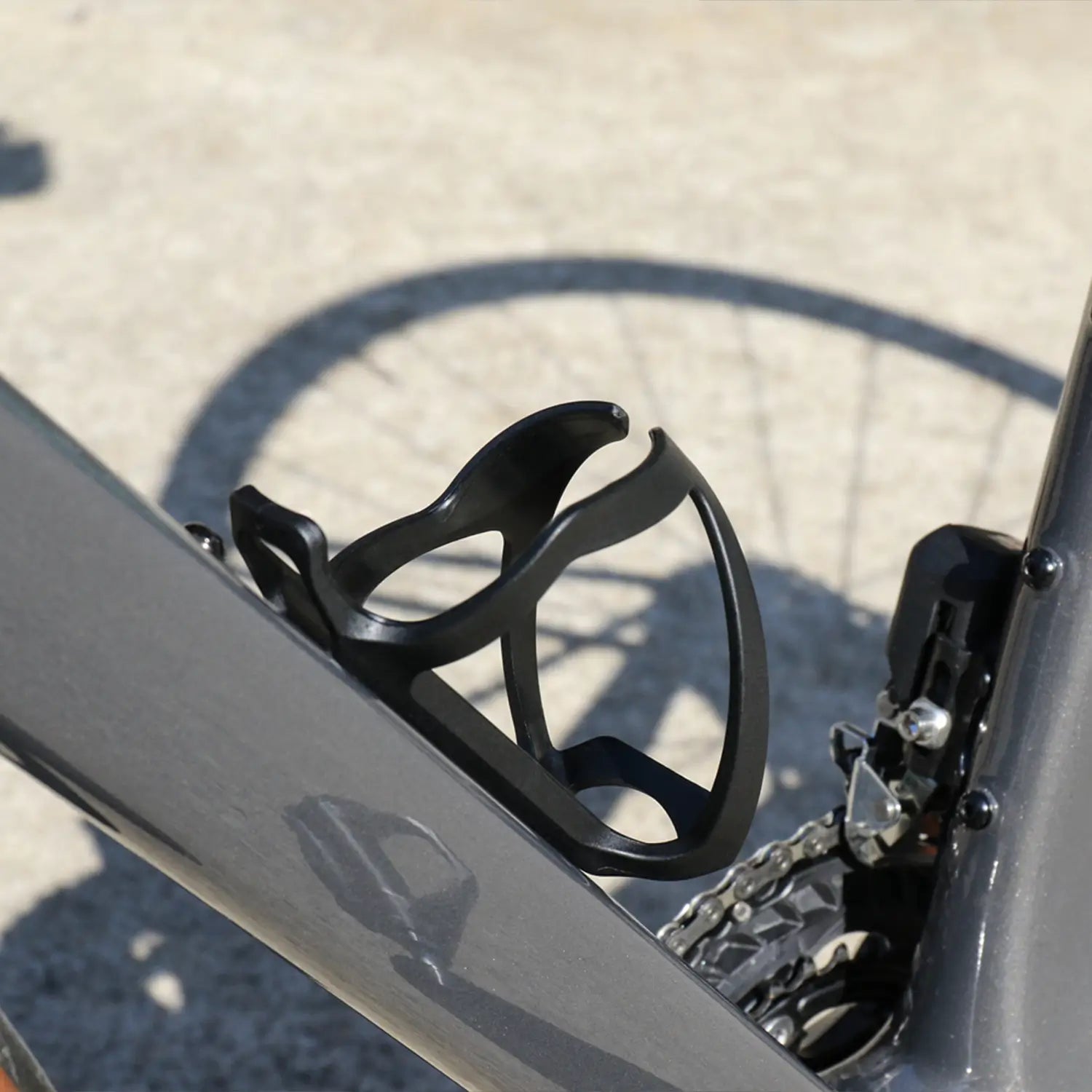 Close-up of a CATEYE BC-500 Bottle Cage, side-loading and black, mounted on a gray bike frame—ideal for bikepacking—with the rear wheel and gear mechanism seen in the background.