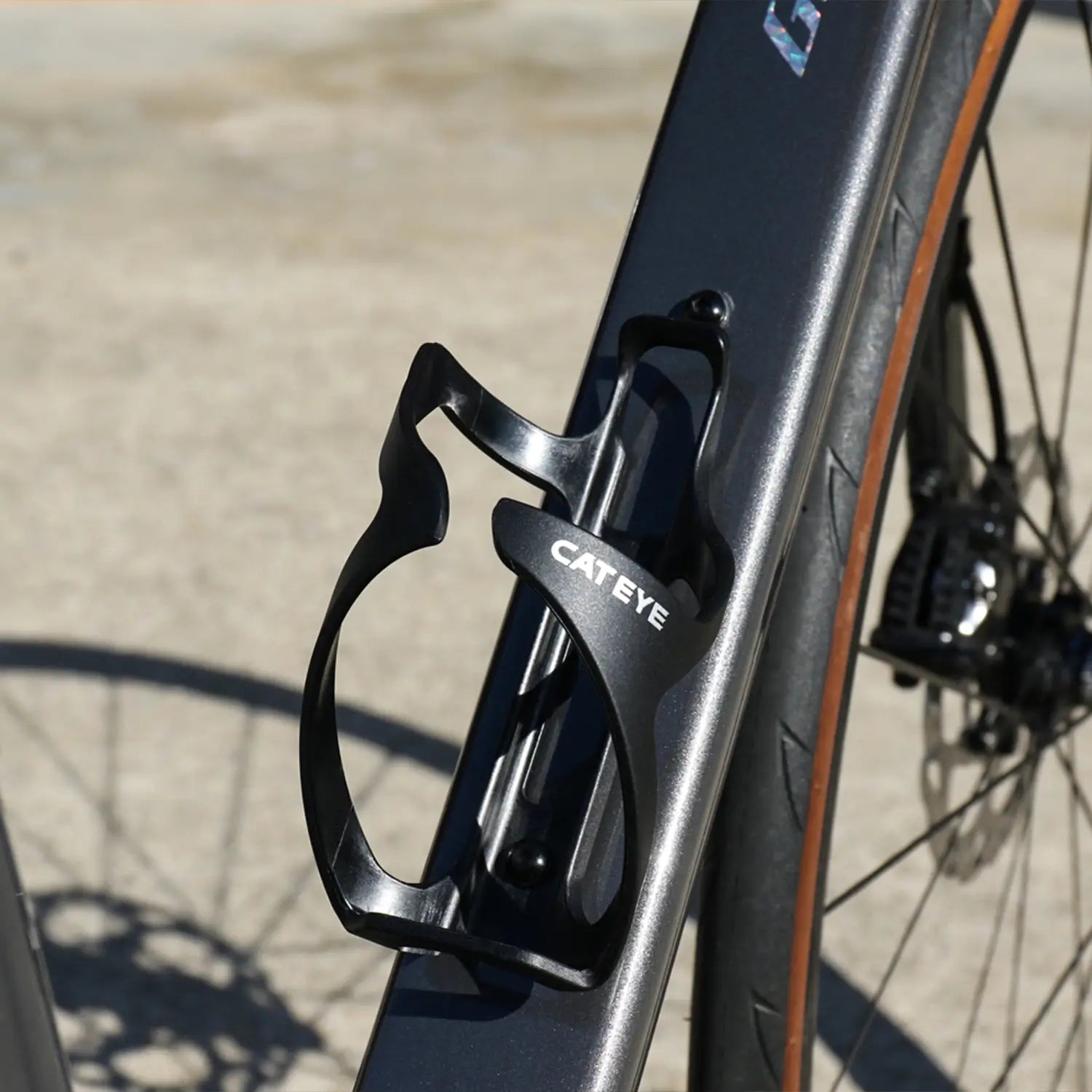 A close-up of the black CATEYE BC-500 Bottle Cage on a matte black bike frame, with the rear wheel and disc brake in the background—ideal for bikepacking adventures.