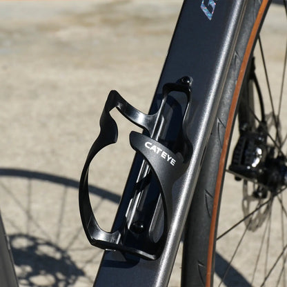 A close-up of the black CATEYE BC-500 Bottle Cage on a matte black bike frame, with the rear wheel and disc brake in the background—ideal for bikepacking adventures.