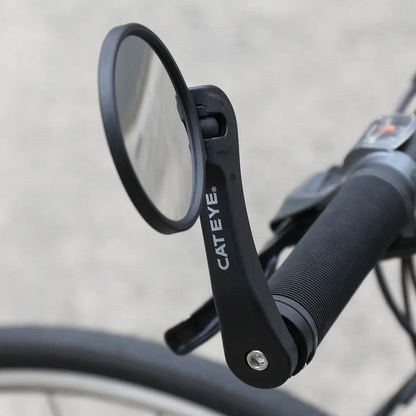 Close-up of a CATEYE BM-600 Handlebar Mirror with a stainless steel lens attached to a black bike handlebar, providing enhanced rear visibility against a blurred pavement background.