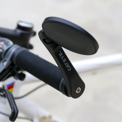 Close-up of a CATEYE BM-600 Handlebar Mirror with a stainless steel lens, mounted on a bike handlebar for enhanced rear visibility. Part of the brake lever and frame are visible in the background.
