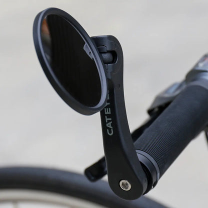A close-up of a CATEYE BM-600 Handlebar Mirror with a stainless steel lens on a bicycle handlebar, showing the grip, part of the brake lever, and a blurred front wheel for better rear visibility.