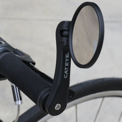 A close-up of a bicycle handlebar with the CATEYE BM-600 Handlebar Mirror, which has a stainless steel lens for better rear visibility. Part of the bike’s rear wheel is visible in the background.