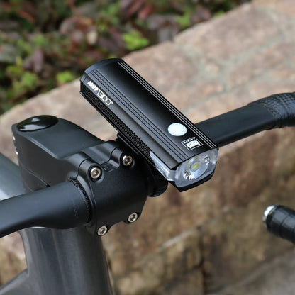 A black CATEYE AMPP1300 bicycle headlight with OptiCube lens technology is mounted on bike handlebars, with a stone wall and foliage blurred in the background.