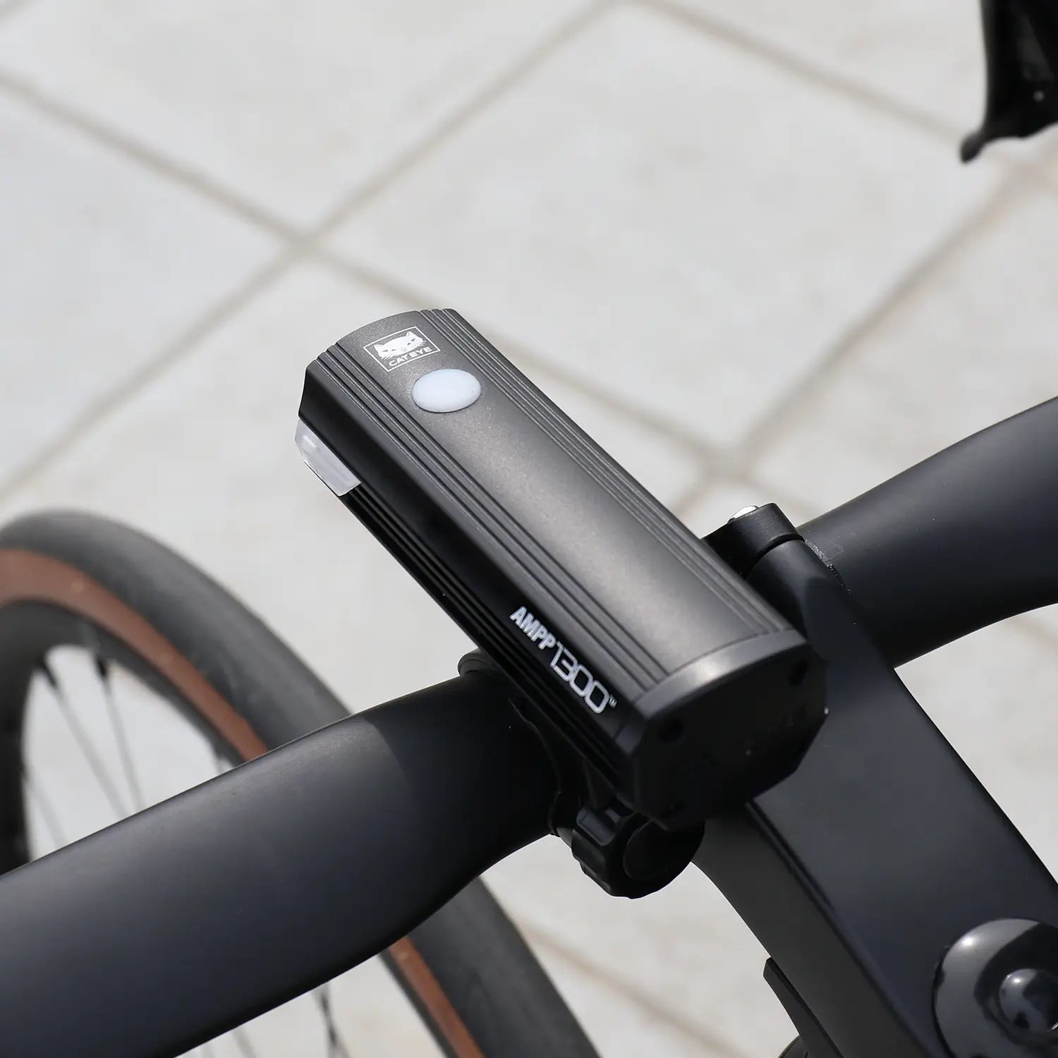 A close-up of the CATEYE AMPP1300 bike headlight with OptiCube™ lens technology, mounted on handlebars using a FlexTight™ bracket. The front tire and paved ground are partially visible in the background.