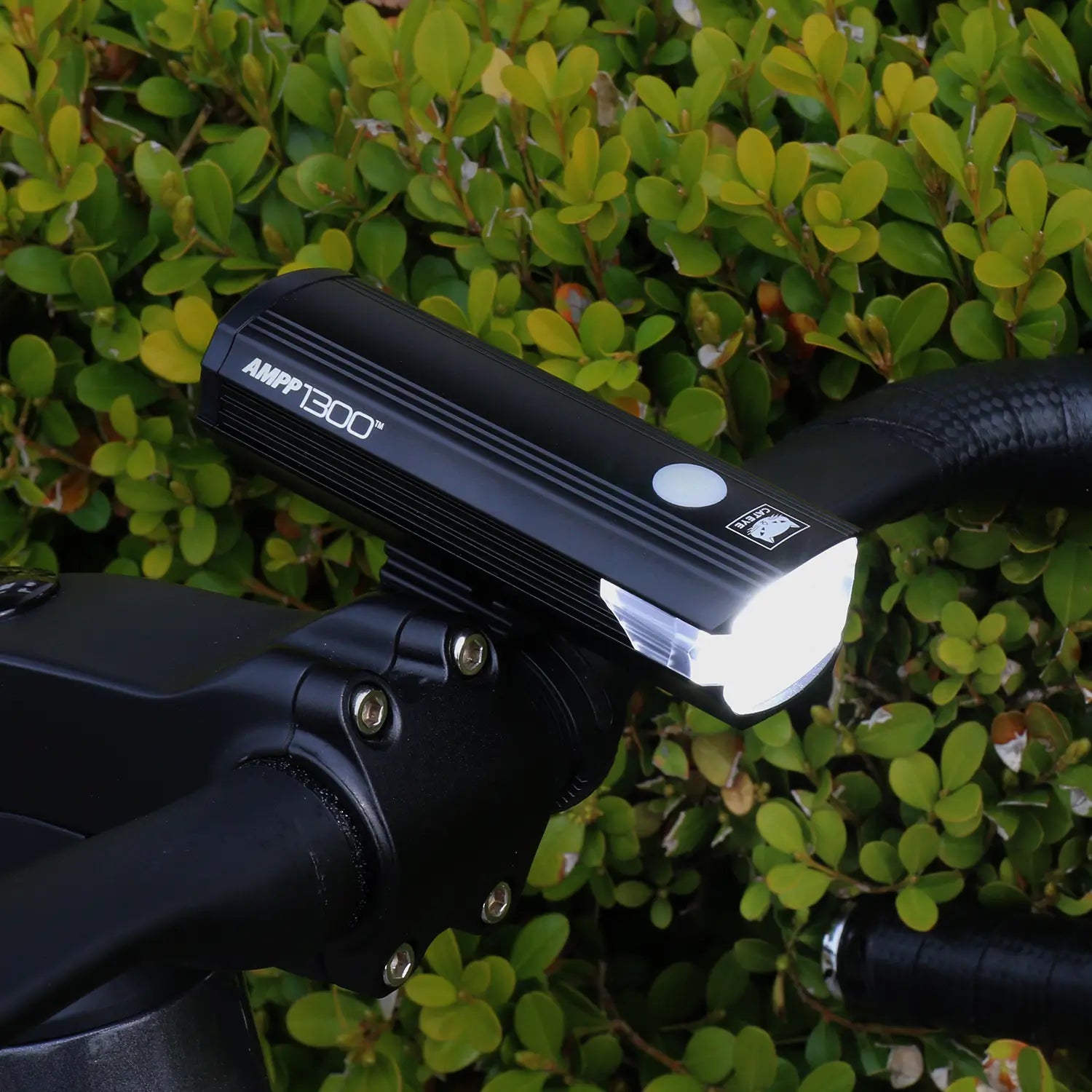 A CATEYE AMPP1300 bicycle headlight with OptiCube lens technology is mounted on a handlebar, emitting a bright white light against green leafy bushes in the background.