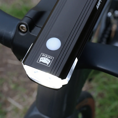 Close-up of a black CATEYE AMPP1300 bicycle headlight with OptiCube lens technology, mounted on handlebars and shining a bright white light. Blurred grass and part of a bike wheel are visible in the background.