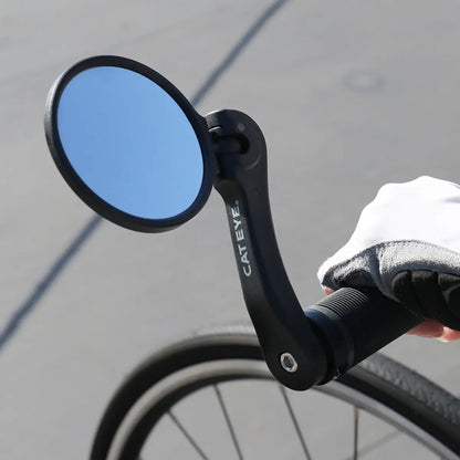 A close-up of the CATEYE BM-600 Handlebar Mirror with a stainless steel lens attached to a bike handlebar, showing a gloved hand gripping the bar and part of the wheel in view for improved rear visibility.