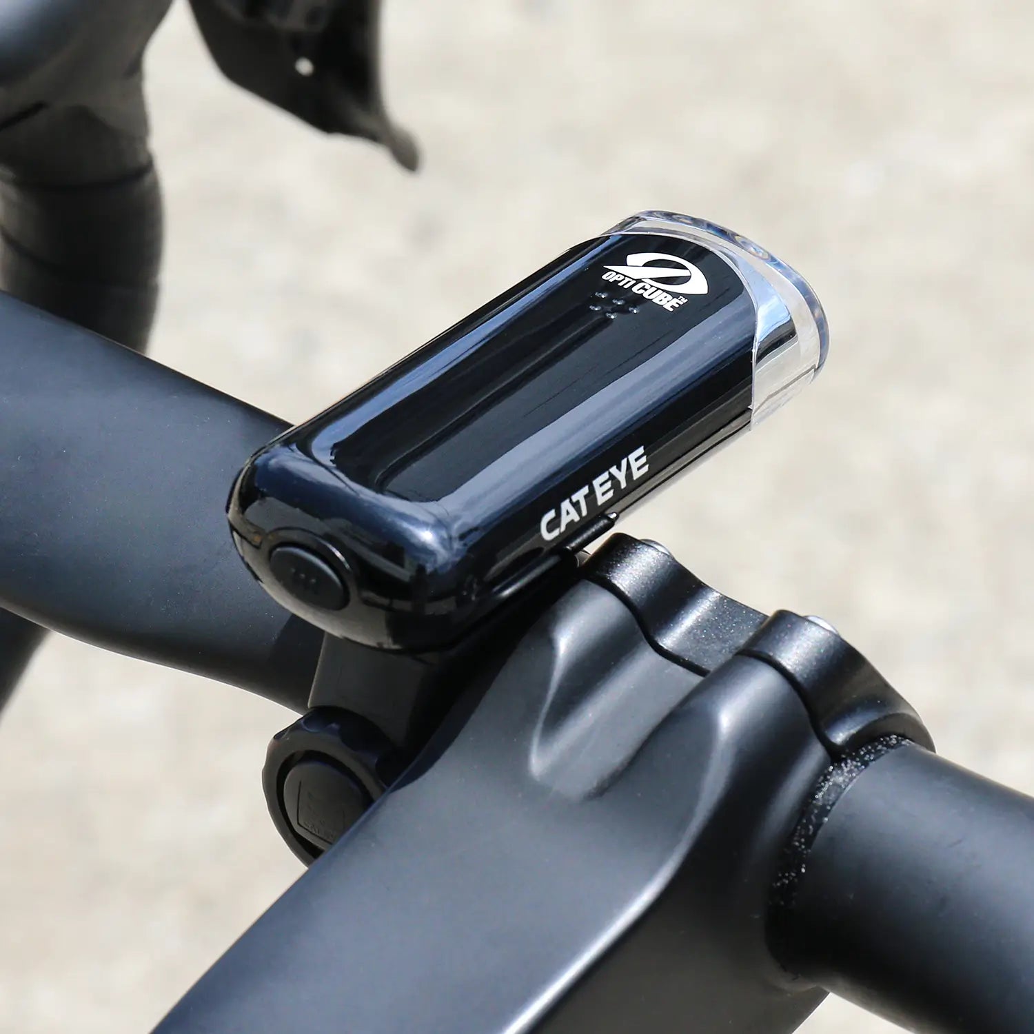 A CATEYE Hl-El135N black bicycle headlight with an OptiCube™ lens is mounted on the handlebars, set against a blurred outdoor background.