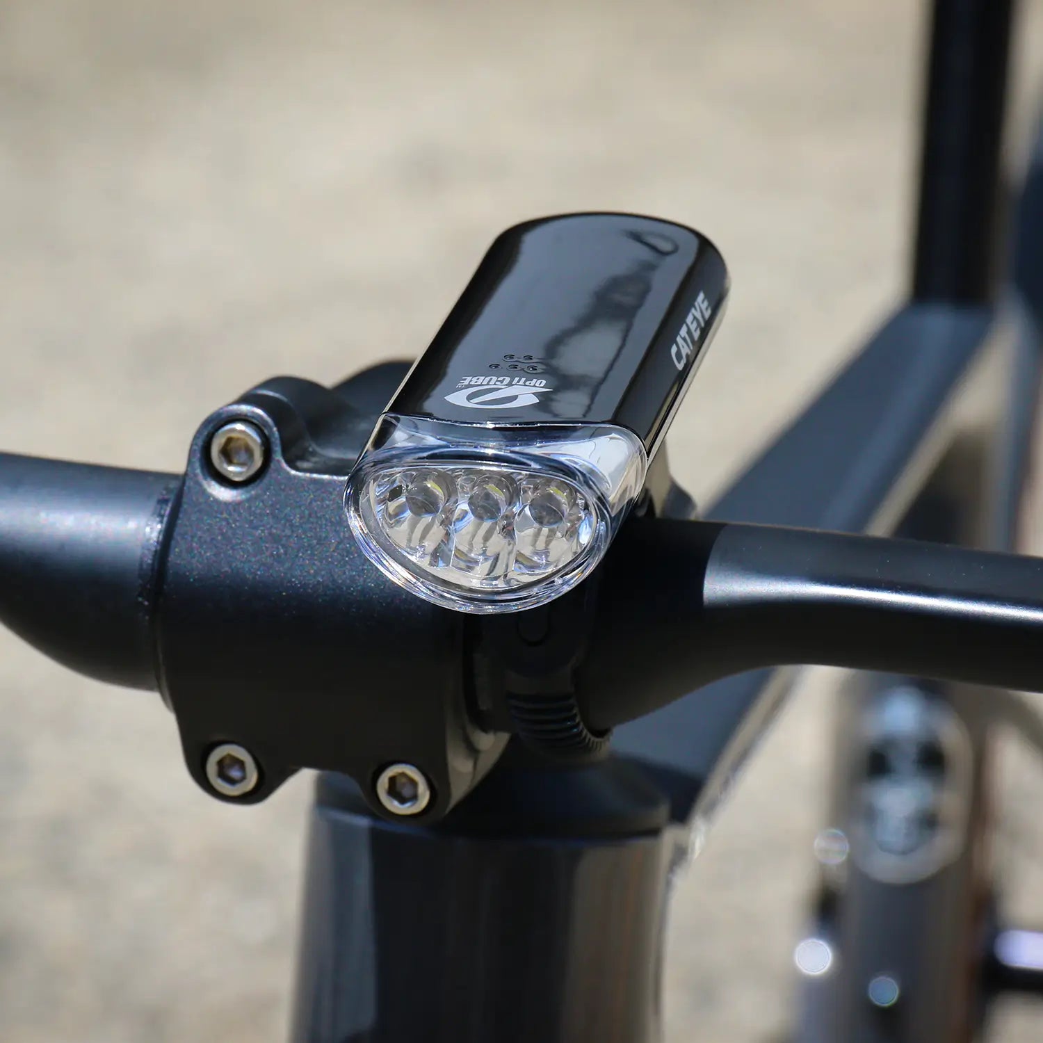 A close-up of a black bicycle handlebar with the CATEYE Hl-El135N front bike safety light, featuring three LED bulbs and an OptiCube™ lens, mounted outdoors in daylight.