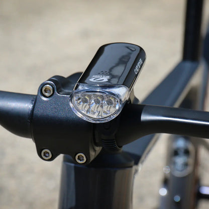 A close-up of a black bicycle handlebar with the CATEYE Hl-El135N front bike safety light, featuring three LED bulbs and an OptiCube™ lens, mounted outdoors in daylight.