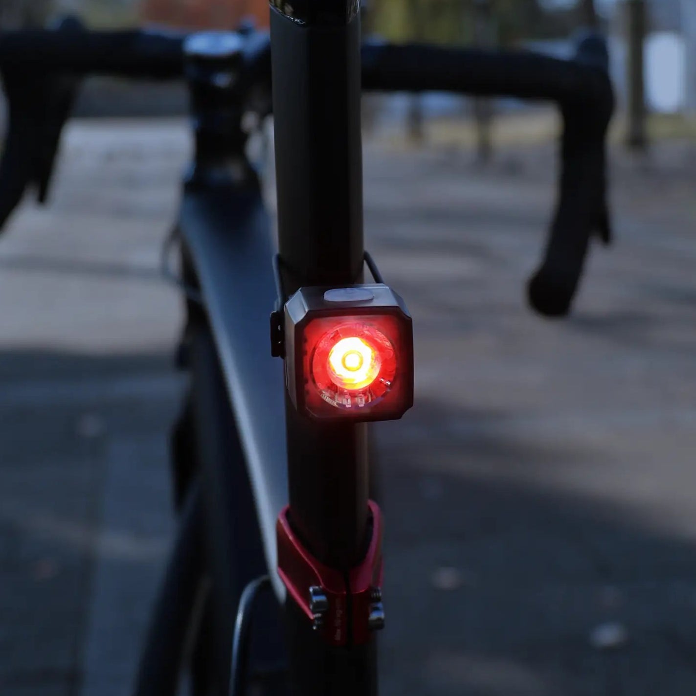 Close-up of the CATEYE NANO60 USB-C rechargeable bike light illuminated on a bicycle’s rear, outdoors on a paved path with blurred trees and buildings in the background.