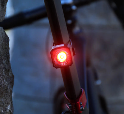 A close-up of the CATEYE AMPP400S & NANO60 red LED bike light glowing brightly on a seat post, with a stone wall and blurred bike frame in the background.