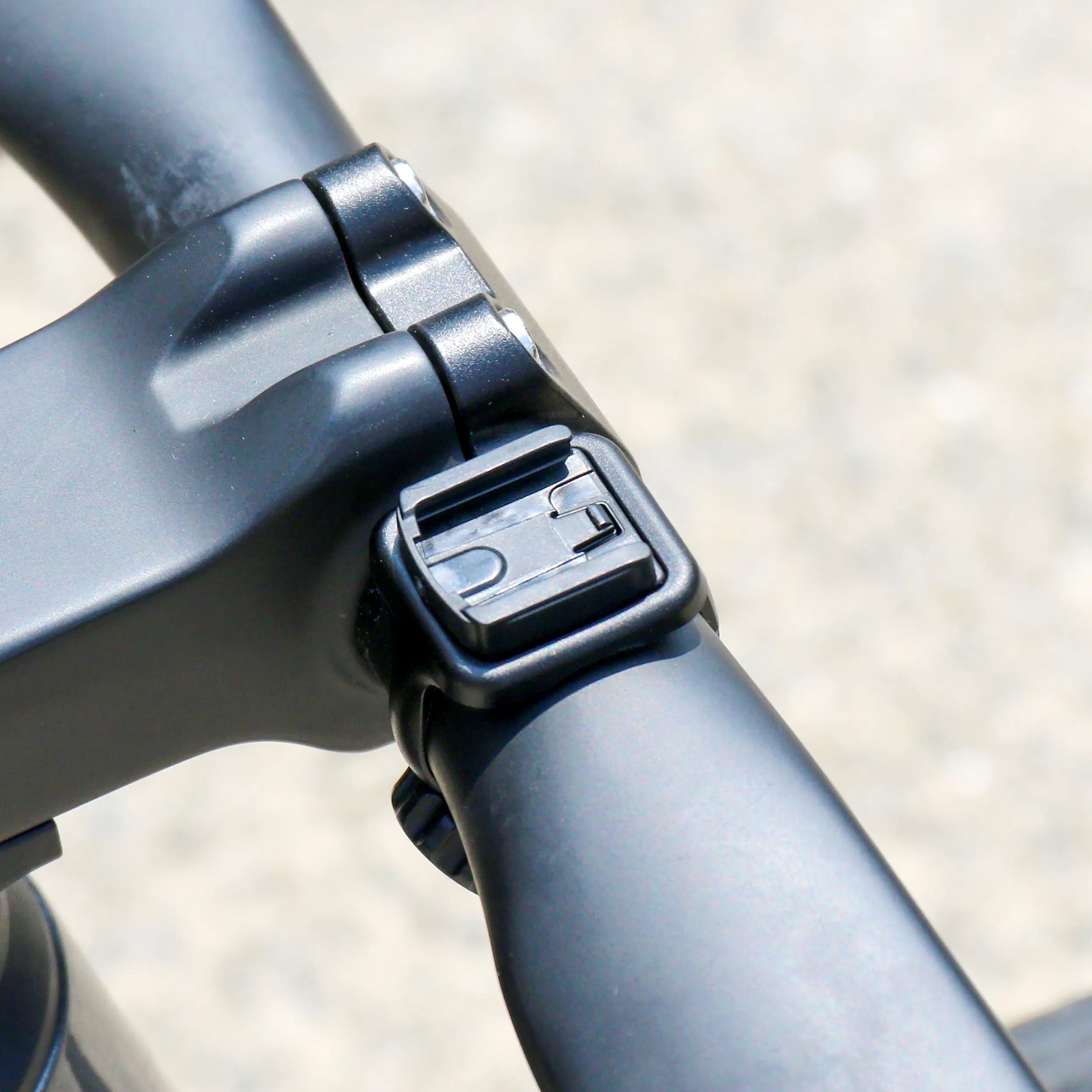 Close-up of a black handlebar and stem joint with a small lever, featuring the CATEYE Flextight™ Computer Mount, compatible with CatEye wireless cycling computers, shown outdoors in bright sunlight against a blurred background.