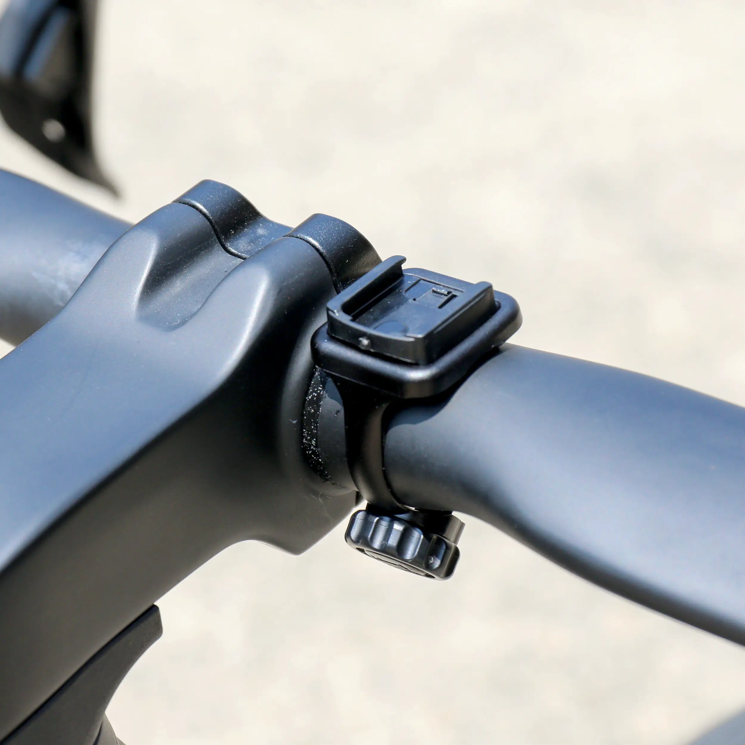 Close-up of a black bicycle handlebar with a CATEYE Flextight™ Computer Mount attached near the stem using a strap and tightening knob, perfect for wireless cycling computers, set against a blurred outdoor background.
